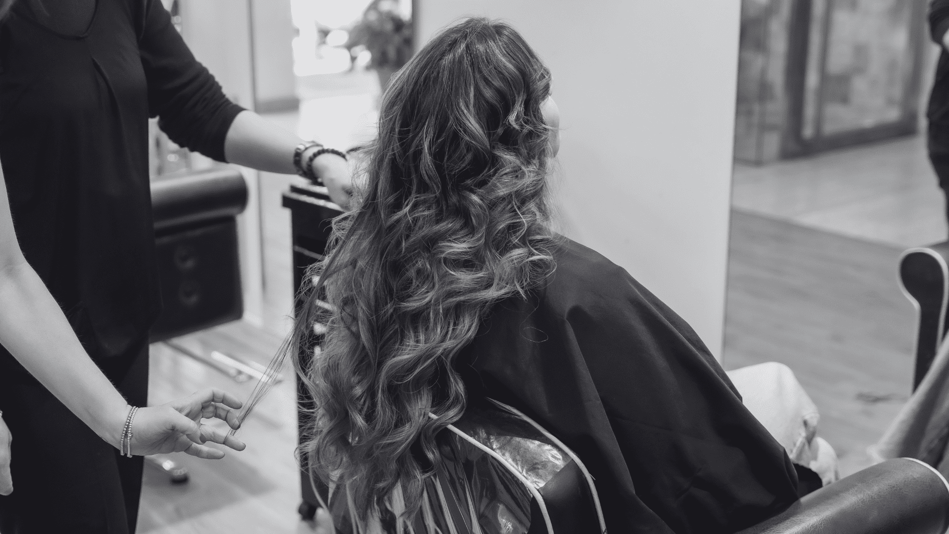 Hair stylist curling long hair in salon chair, with mirrors and styling tools in the background.