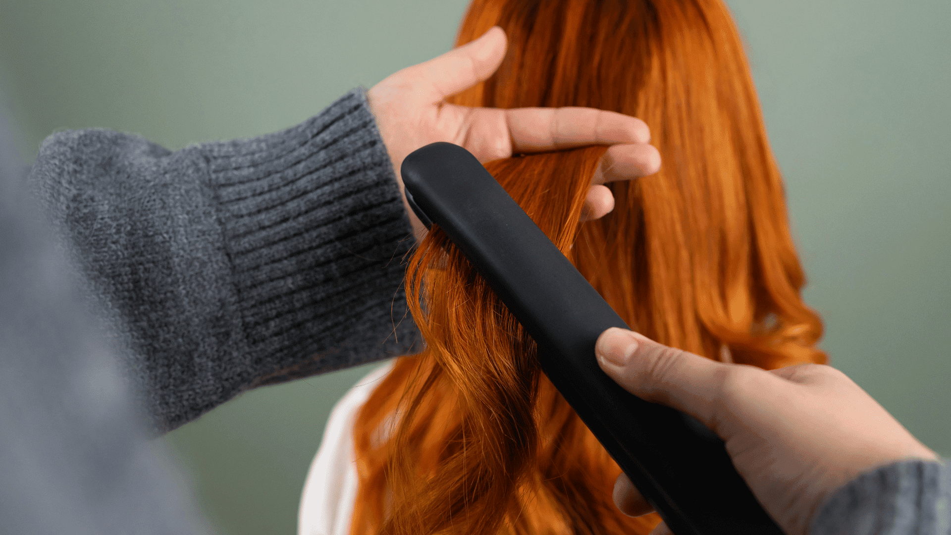 Hair stylist using a flat iron on red hair against a green background.