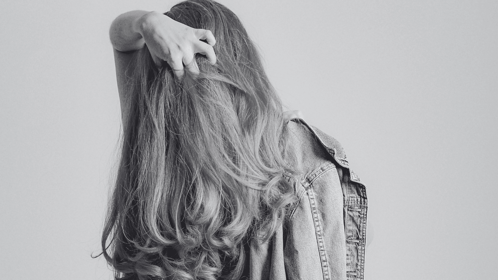 A woman with long curly hair stands with her back to the camera, hand in her hair, wearing a jacket.