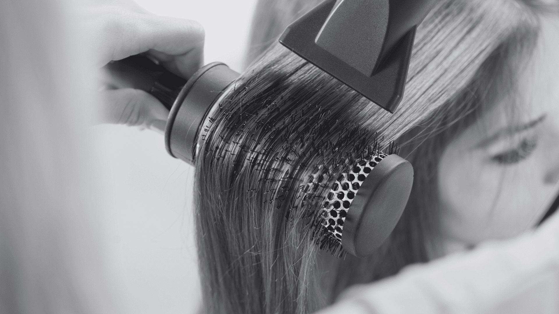 Hair stylist using a round brush and hairdryer to style client's long, straight hair.